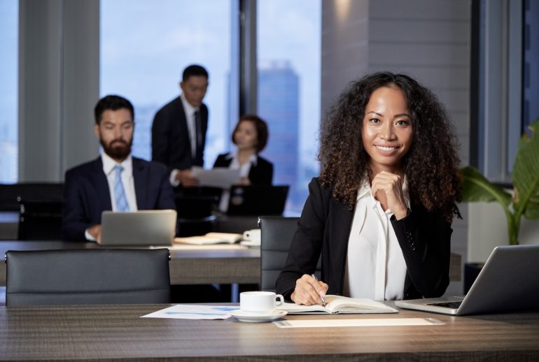 Beautiful ethnic formal woman sitting at table with laptop and papers smiling at camera with coworkers on background